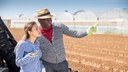 Young Colombian woman and African American man farmers talking outdoors on background with ploughed field and industrial greenhouses

